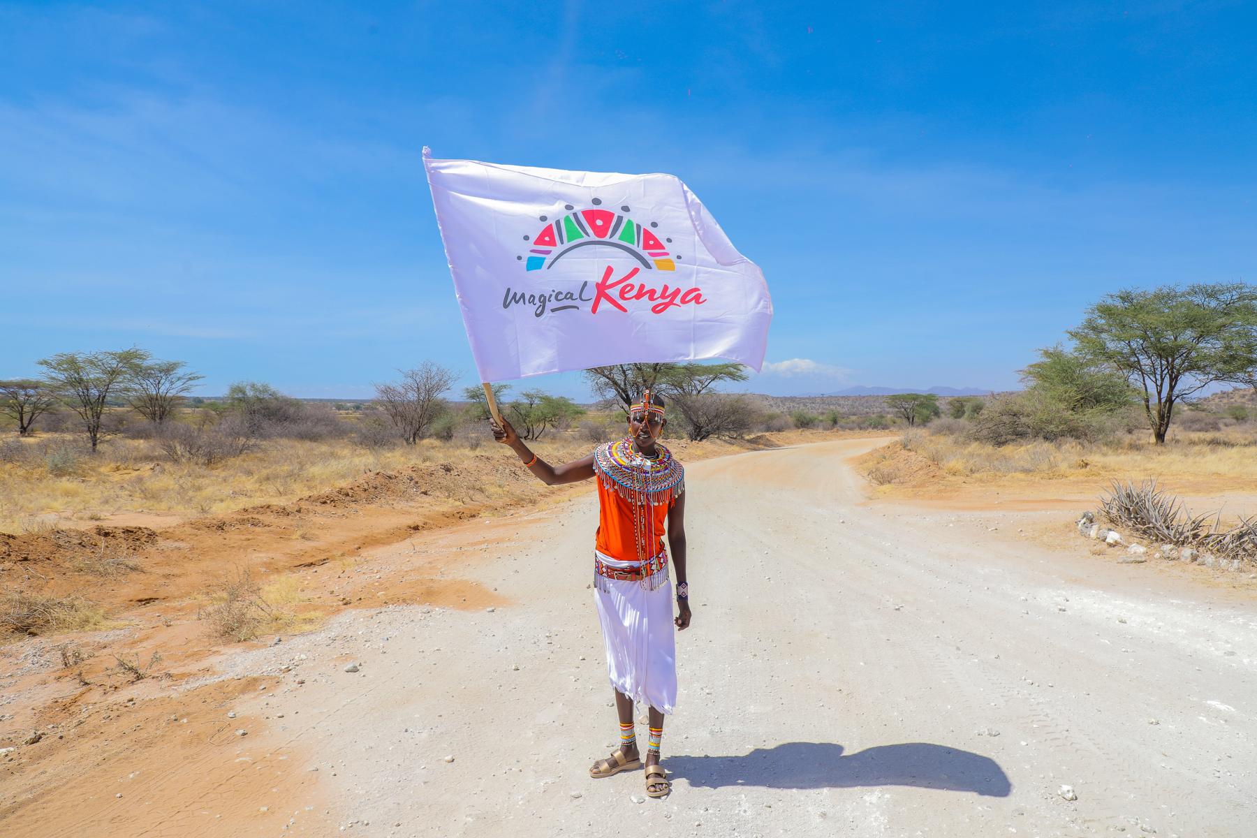 A Samburu woman in beaded jewellery waves a Magical Kenya flag on a dusty road