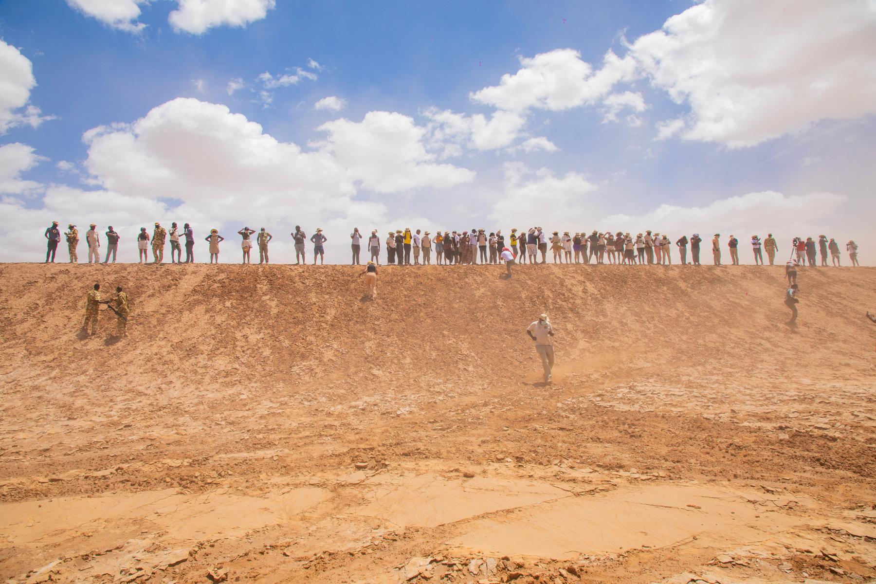A long line of travellers stands along the top of a dune ridge while others climb its sandy face