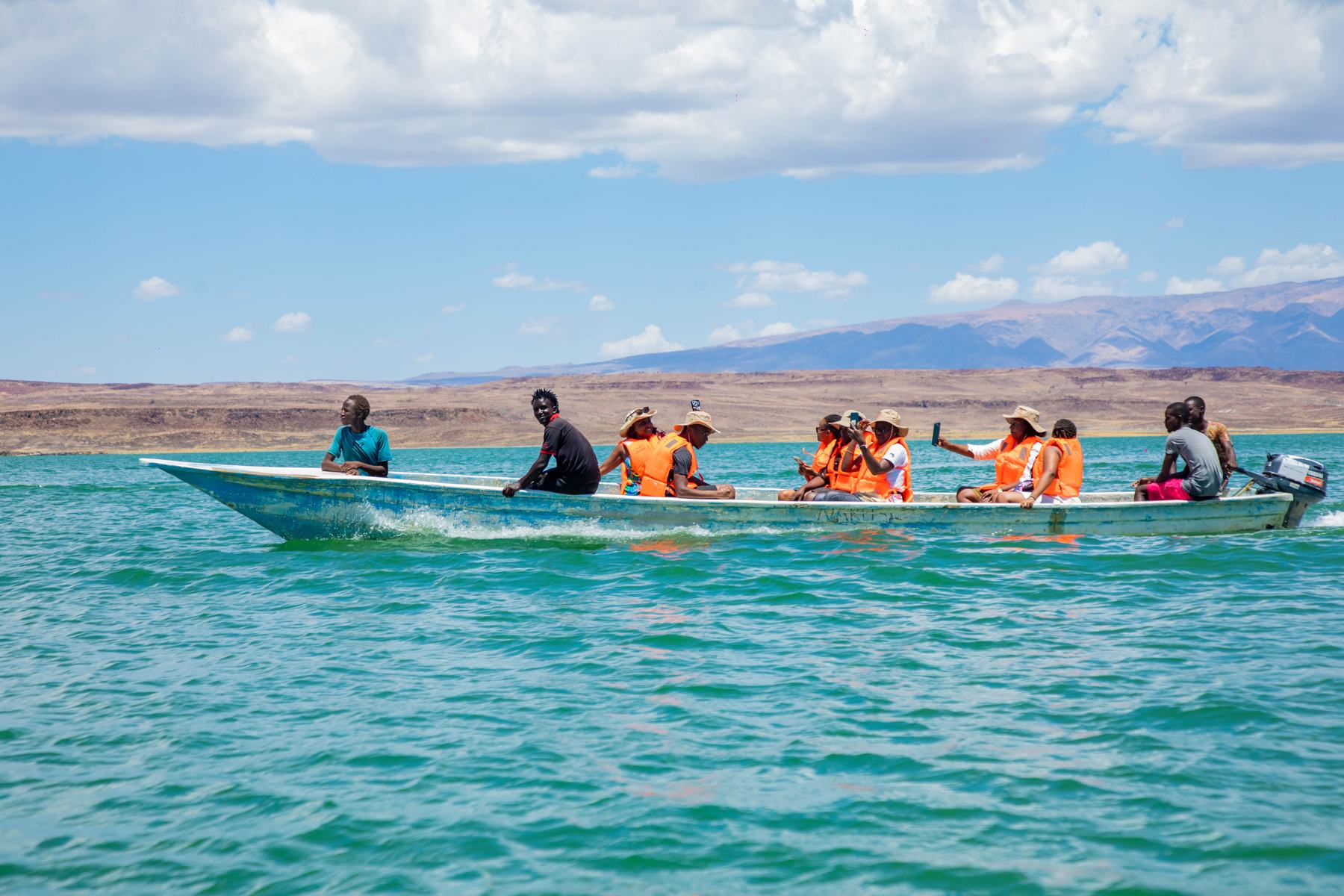 Travellers in orange life jackets ride a long boat across the turquoise water of Lake Turkana, desert hills behind