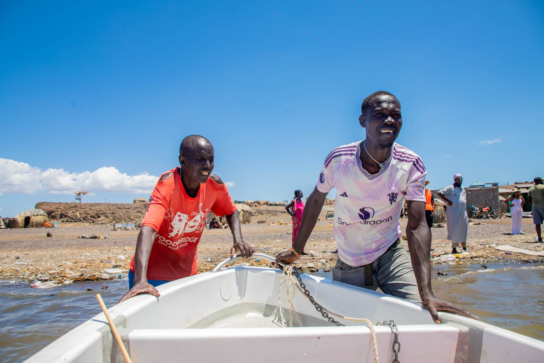 Two smiling fishermen push a white boat out from the shore of Lake Turkana